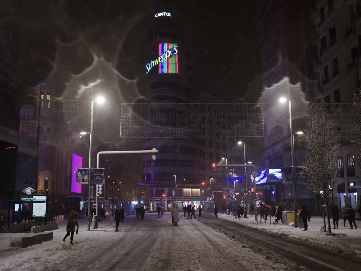 Gran Vía in central Madrid lit up at night during a rare snowfall, with pedestrians crossing the snow-covered street