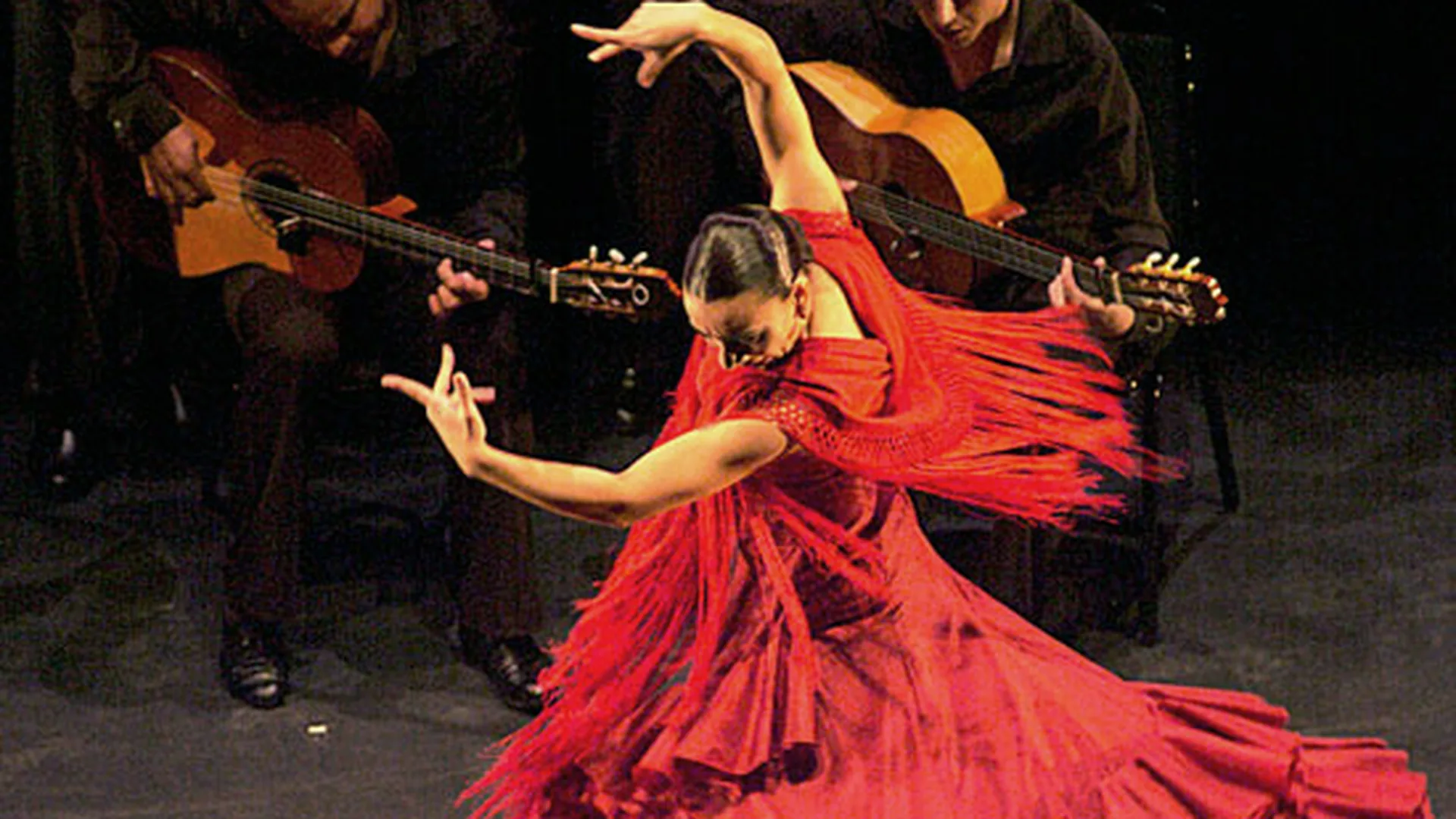 A flamenco dancer in a red dress mid-performance on a small stage, spotlit, with a guitarist seated behind her